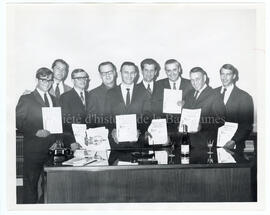 9 hommes [inconnus] posant avec des feuilles dans les mains. De gauche à droite, on aperçoit Guy ...